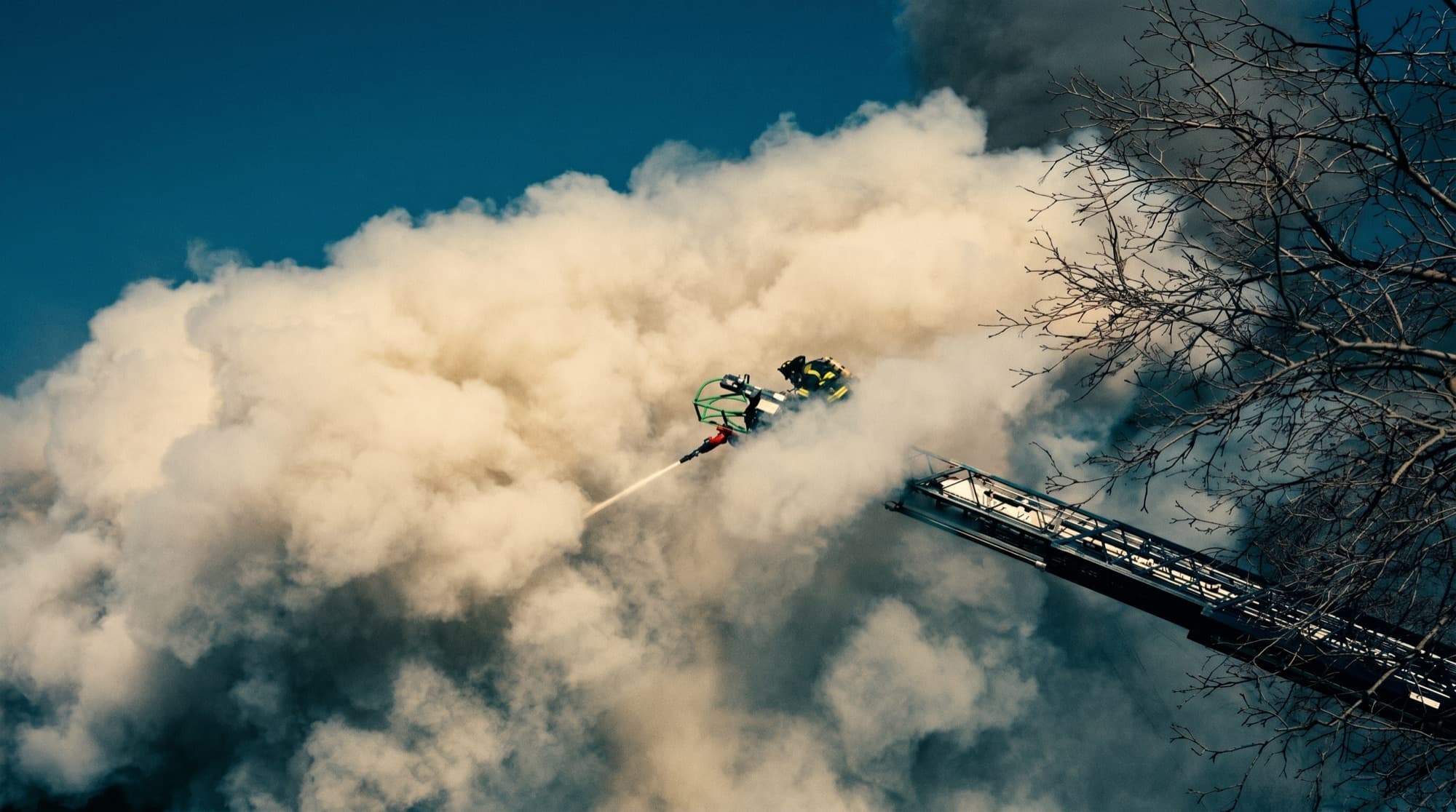 A firefighter works from an aerial ladder platform, directing a hose stream into heavy smoke.