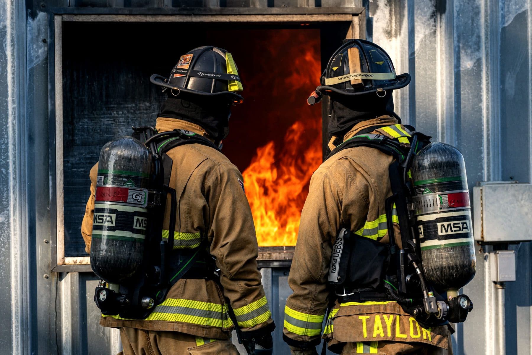 Two firefighters facing an open burn-cell window during a live training burn.